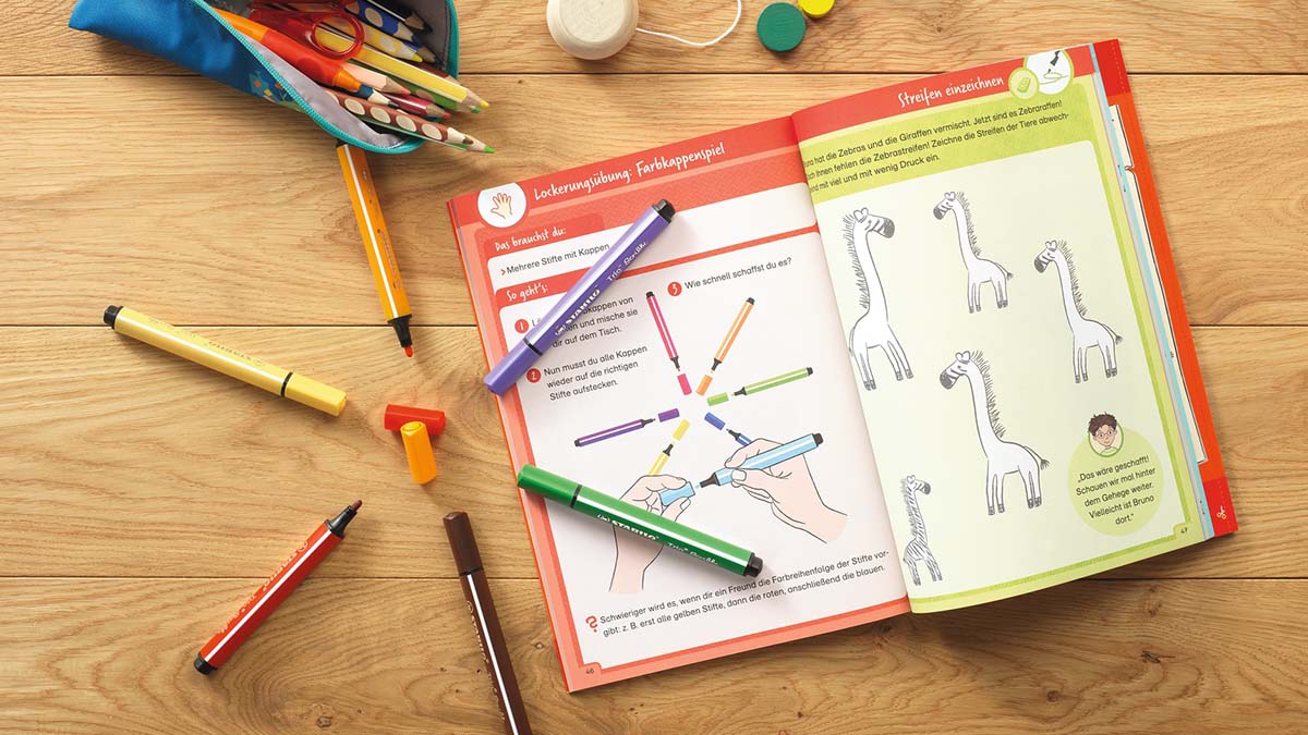 Offenes Buch mit Malanleitung und bunten Stiften auf Tisch, kreative Lernumgebung
