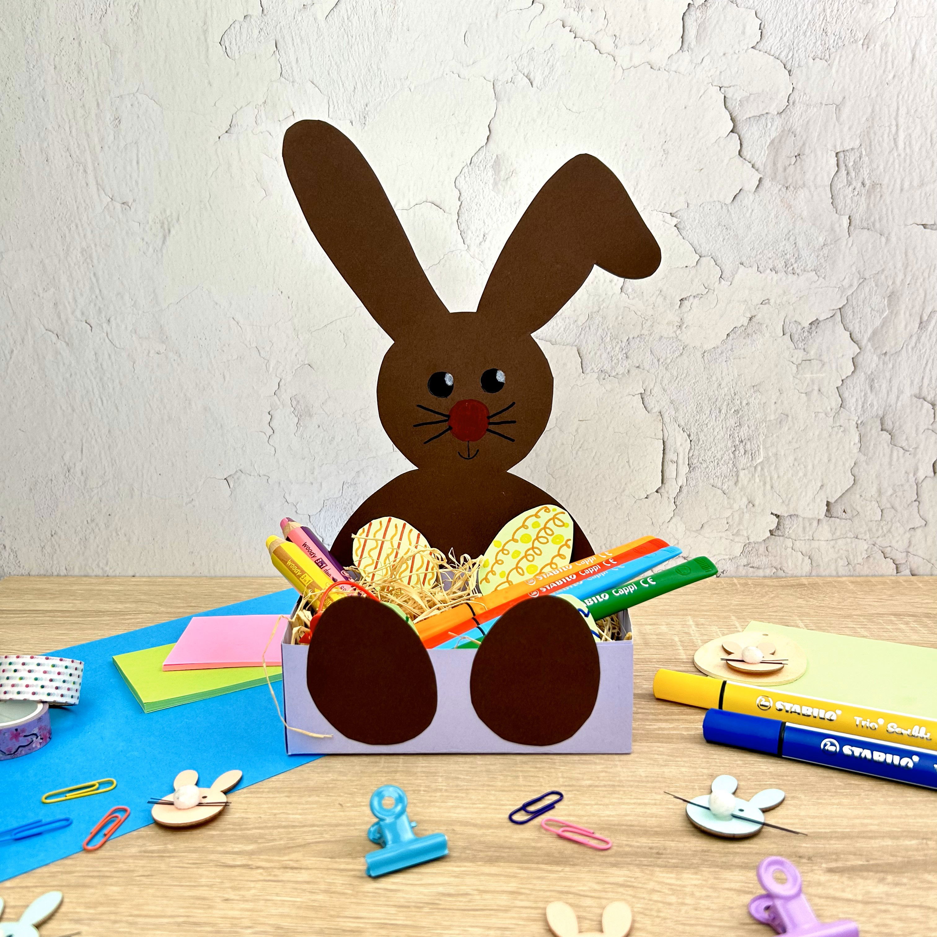 Brauner Papierhase mit bunten Stiften in einem Kasten, umgeben von Bastelmaterialien.