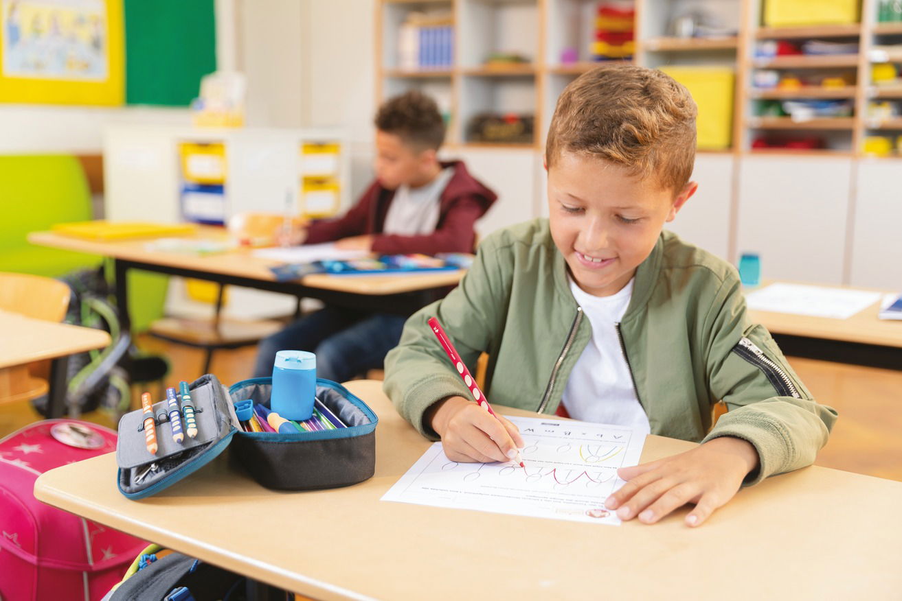 Schüler schreibt konzentriert an einem Tisch in einem Klassenzimmer, mit Arbeitsblatt.